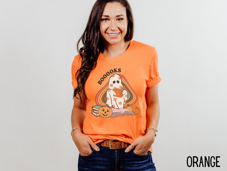 May include: Orange t-shirt featuring a ghost reading a book with the text "BOO-OOOKS" in a rainbow arch. The ghost is wearing a floral pattern and there are pumpkins and books on the ground.