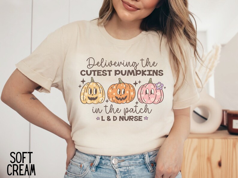 May include: Soft cream-colored t-shirt with the text "Delivering the CUTEST PUMPKINS in the patch L & D NURSE" and three cartoon pumpkins. The pumpkins are orange, yellow, and pink with smiling faces. The shirt is a casual style.