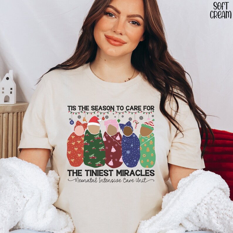 K&ouml;nnte beinhalten: Ein beiges T-Shirt mit einem weihnachtlichen Design, das eine Gruppe von Babys zeigt, die in Decken mit festlichen Mustern gewickelt sind. Der Text lautet "Tis the season to care for the tiniest miracles - Neonatal Intensive Care Unit".