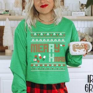 K&ouml;nnte beinhalten: Gr&uuml;ner Weihnachtspullover mit einem festlichen Muster und dem Text "Merry Xmas". Der Pullover zeigt eine rote und wei&szlig;e Pille, ein rotes Kreuz und eine wei&szlig;e Krankenschwesterm&uuml;tze.