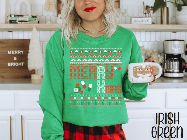 K&ouml;nnte beinhalten: Gr&uuml;ner Weihnachtspullover mit einem festlichen Muster und dem Text "Merry Xmas". Der Pullover zeigt eine rote und wei&szlig;e Pille, ein rotes Kreuz und eine wei&szlig;e Krankenschwesterm&uuml;tze.