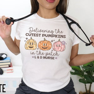 May include: White t-shirt with a colorful graphic of three pumpkins and the text "Delivering the cutest pumpkins in the patch L&D Nurse." A black stethoscope is draped around the neck. The word "WHITE" is at the bottom.