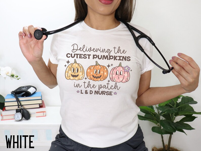 May include: White t-shirt with a colorful graphic of three pumpkins and the text "Delivering the cutest pumpkins in the patch L&D Nurse." A black stethoscope is draped around the neck. The word "WHITE" is at the bottom.
