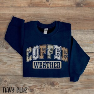 May include: Navy blue sweatshirt with the words "COFFEE WEATHER" in a distressed, vintage-style font. The sweatshirt is made of a soft material and is displayed on a wooden surface. The words "NAVY BLUE" are at the bottom.