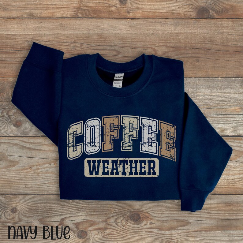 May include: Navy blue sweatshirt with the words "COFFEE WEATHER" in a distressed, vintage-style font. The sweatshirt is made of a soft material and is displayed on a wooden surface. The words "NAVY BLUE" are at the bottom.