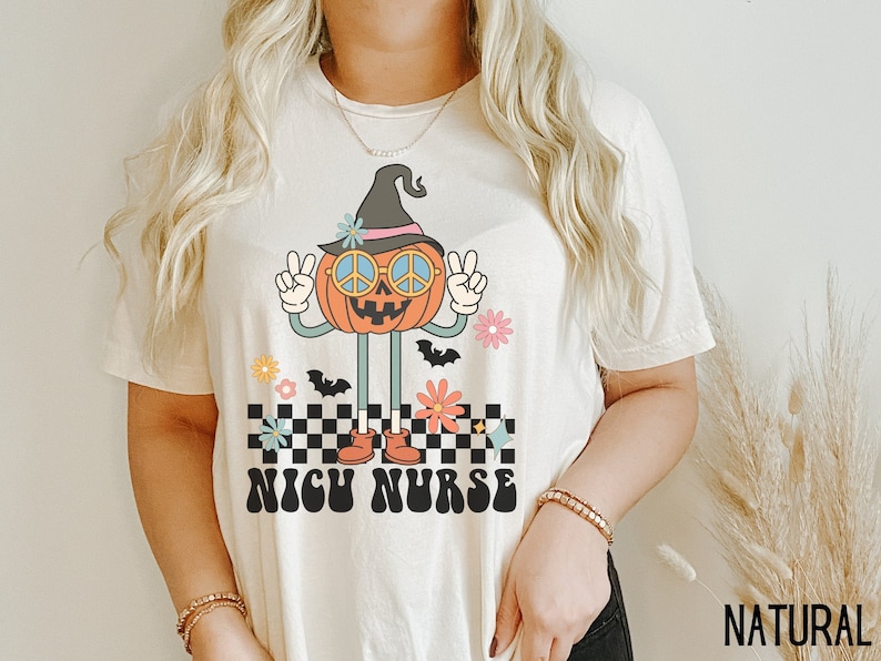Puede incluir: Una camiseta blanca con un gr&aacute;fico de una calabaza sonriente que lleva un sombrero de bruja y gafas de sol. La calabaza est&aacute; de pie sobre un patr&oacute;n a cuadros con flores y murci&eacute;lagos. El texto "NICU NURSE" est&aacute; impreso en una fuente retro debajo de la calabaza.