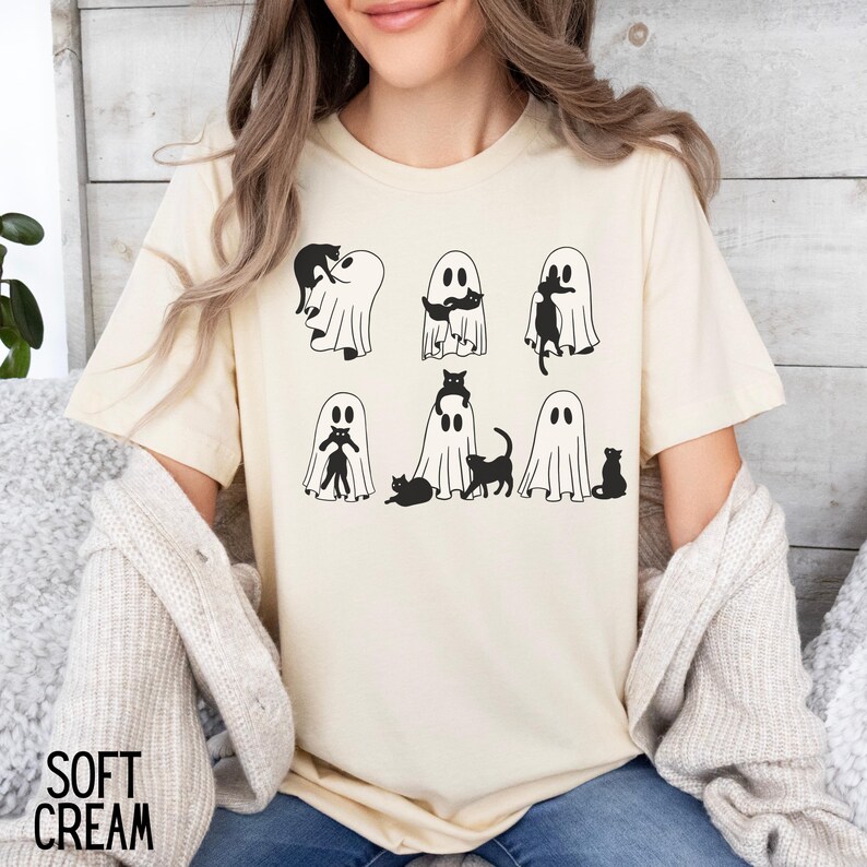 Puede incluir: Una camiseta blanca con un gr&aacute;fico negro de gatos y fantasmas. El gr&aacute;fico muestra seis fantasmas con gatos dentro, fuera y encima de ellos.