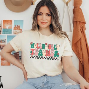 May include: A cream colored t-shirt with a retro-style graphic that says "Holly Jolly Teacher" in colorful letters.