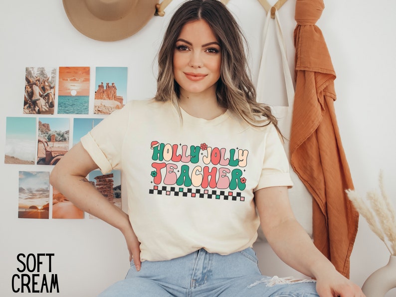 May include: A cream colored t-shirt with a retro-style graphic that says "Holly Jolly Teacher" in colorful letters.