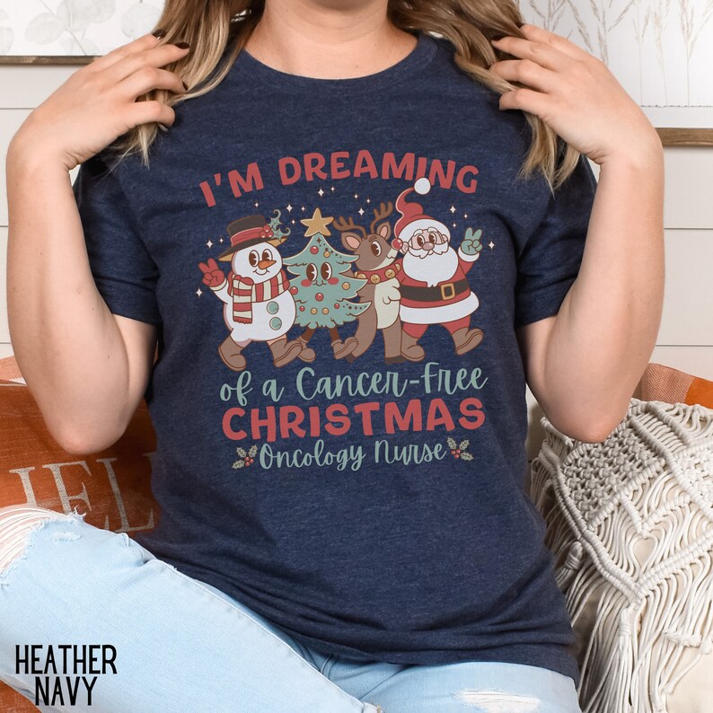 May include: A navy blue t-shirt with a Christmas-themed design featuring a snowman, reindeer, and Santa Claus. The text on the shirt reads "I'm Dreaming of a Cancer-Free Christmas Oncology Nurse".