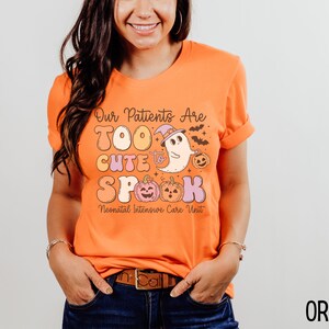 May include: Orange t-shirt with a Halloween-themed graphic that says "Our Patients Are Too Cute To Spook Neonatal Intensive Care Unit". The graphic features a ghost wearing a witch hat and holding a pumpkin.