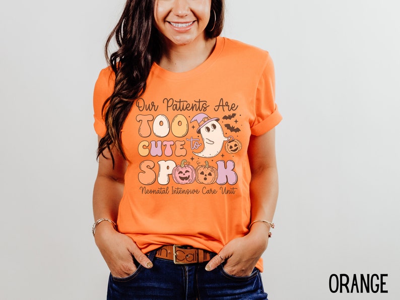May include: Orange t-shirt with a Halloween-themed graphic that says "Our Patients Are Too Cute To Spook Neonatal Intensive Care Unit". The graphic features a ghost wearing a witch hat and holding a pumpkin.