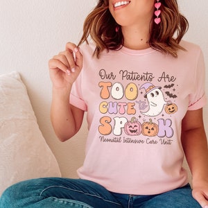 May include: Pink t-shirt with a graphic of a ghost wearing a witch hat and holding a pumpkin. The text on the shirt reads "Our Patients Are Too Cute to Spook Neonatal Intensive Care Unit".