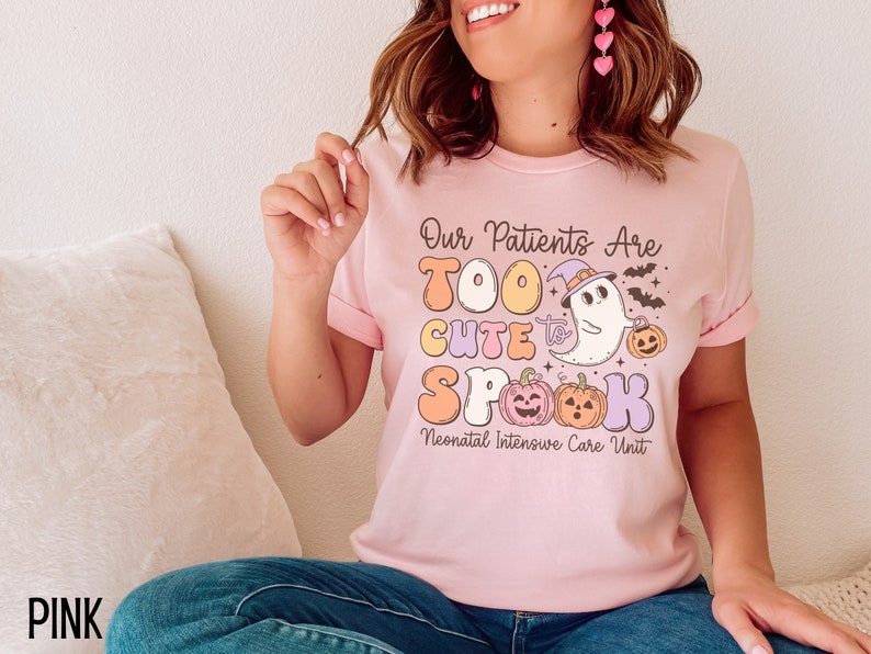 May include: Pink t-shirt with a graphic of a ghost wearing a witch hat and holding a pumpkin. The text on the shirt reads "Our Patients Are Too Cute to Spook Neonatal Intensive Care Unit".