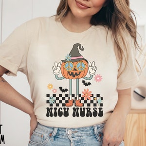Puede incluir: Una camiseta beige con una calabaza de dibujos animados que lleva un sombrero de bruja y gafas de sol con el signo de la paz. La calabaza est&aacute; de pie sobre un patr&oacute;n a cuadros con el texto "NICU NURSE" en una fuente retro.
