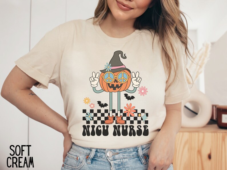Puede incluir: Una camiseta beige con una calabaza de dibujos animados que lleva un sombrero de bruja y gafas de sol con el signo de la paz. La calabaza est&aacute; de pie sobre un patr&oacute;n a cuadros con el texto "NICU NURSE" en una fuente retro.
