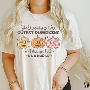 May include: Cream-colored t-shirt with the text "Delivering the Cutest Pumpkins in the Patch" and three cartoon pumpkins in yellow, orange, and pink. The shirt also includes the text "L & D Nurse".