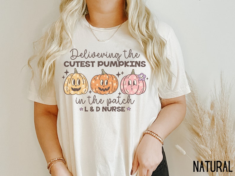 May include: Cream-colored t-shirt with the text "Delivering the Cutest Pumpkins in the Patch" and three cartoon pumpkins in yellow, orange, and pink. The shirt also includes the text "L & D Nurse".