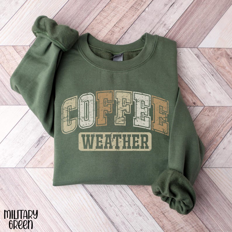 May include: A military green sweatshirt with the words "COFFEE WEATHER" in a vintage, distressed font. The text is in shades of beige and white. The sweatshirt is folded, with the sleeves visible.