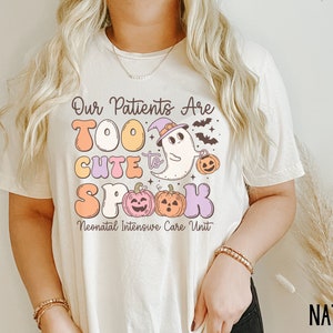 May include: A white t-shirt with a graphic design featuring a ghost wearing a witch hat and holding a pumpkin. The text reads "Our Patients Are Too Cute to Spook Neonatal Intensive Care Unit".