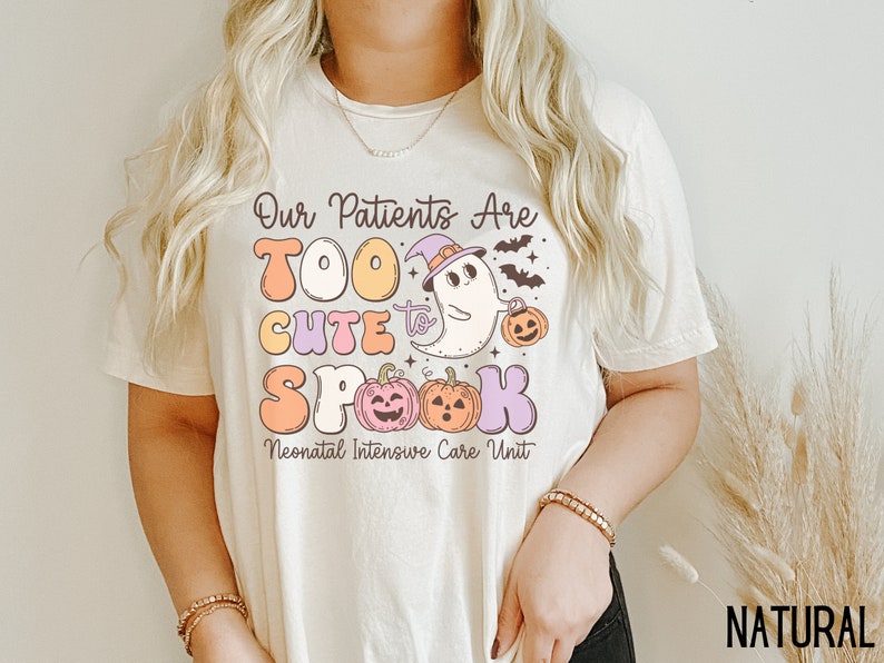 May include: A white t-shirt with a graphic design featuring a ghost wearing a witch hat and holding a pumpkin. The text reads "Our Patients Are Too Cute to Spook Neonatal Intensive Care Unit".