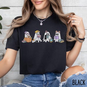 May include: Black t-shirt featuring a whimsical illustration of cartoon ghosts reading books. The ghosts are adorned with accessories like glasses, flowers, and a pumpkin. The word "BLACK" is printed at the bottom of the shirt.
