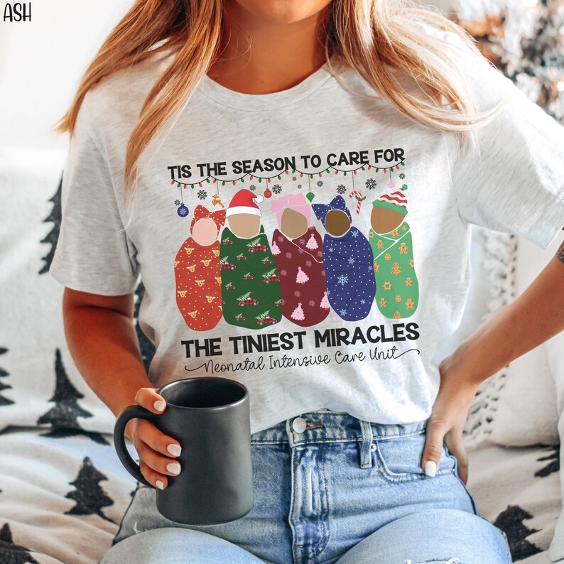K&ouml;nnte beinhalten: Ein wei&szlig;es T-Shirt mit einem weihnachtlichen Design, das f&uuml;nf Babys zeigt, die in Decken mit verschiedenen Weihnachtsmustern gewickelt sind. Der Text lautet "Tis the season to care for the tiniest miracles - Neonatal Intensive Care Unit".