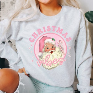 May include: A light gray sweatshirt with a pink and white Christmas design. The design features a cartoon Santa Claus with a candy cane and the text "Christmas Vibes".
