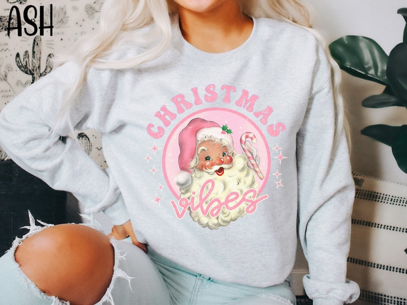 May include: A light gray sweatshirt with a pink and white Christmas design. The design features a cartoon Santa Claus with a candy cane and the text "Christmas Vibes".