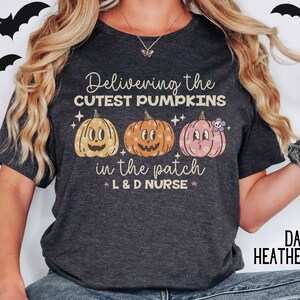 May include: Dark heather gray t-shirt with the text "Delivering the CUTEST PUMPKINS in the patch L & D NURSE" and three cartoon pumpkins. The pumpkins are in shades of orange, yellow, and pink. Black bat silhouettes are in the background.