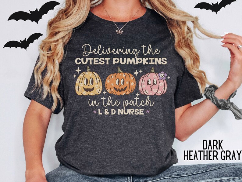 May include: Dark heather gray t-shirt with the text "Delivering the CUTEST PUMPKINS in the patch L & D NURSE" and three cartoon pumpkins. The pumpkins are in shades of orange, yellow, and pink. Black bat silhouettes are in the background.