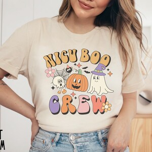 May include: A cream colored t-shirt with a retro-style graphic that says "NIGH BOO CREW" with a pumpkin, ghost, and spider illustration.