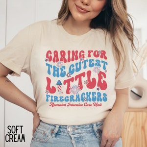May include: A beige t-shirt with a red, white, and blue graphic that says "Caring for the Cutest Little Firecrackers Neonatal Intensive Care Unit".