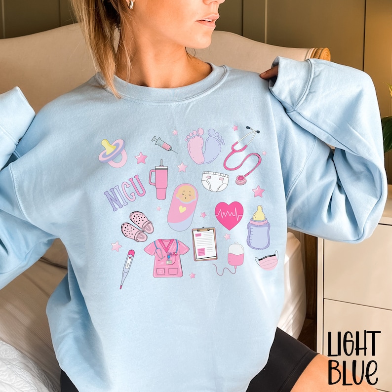 May include: Light blue sweatshirt with a graphic design of various medical symbols and the word "NICU". The symbols include a stethoscope, a syringe, a baby bottle, a pacifier, a baby footprint, a heart rate monitor, a thermometer, a diaper, a baby in a crib, a medical mask, and a nurse's uniform.