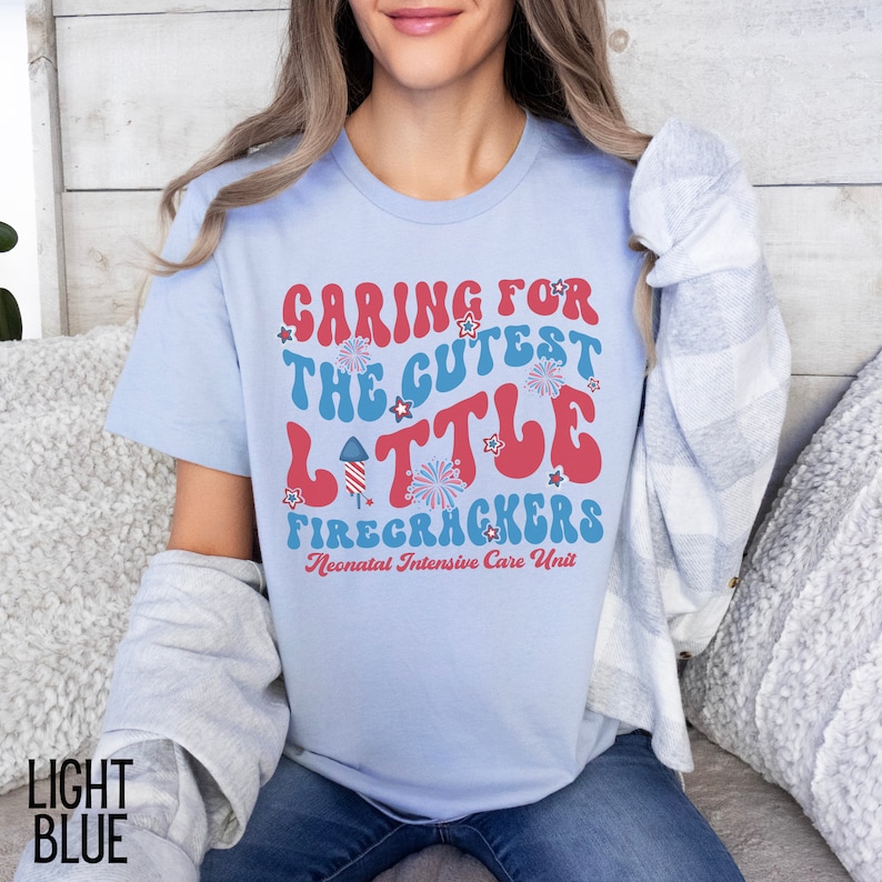 May include: Light blue t-shirt with a red, white, and blue graphic that says "Caring for the Cutest Little Firecrackers" and "Neonatal Intensive Care Unit".