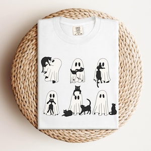 May include: White t-shirt with a black and white graphic of ghosts and cats. The graphic shows eight ghosts with cats in various positions, including a cat riding on a ghost's back, a cat sitting on a ghost's head, and a cat peeking out from behind a ghost.
