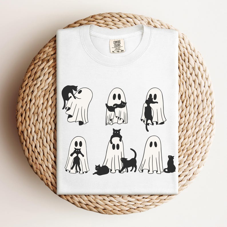 May include: White t-shirt with a black and white graphic of ghosts and cats. The graphic shows eight ghosts with cats in various positions, including a cat riding on a ghost's back, a cat sitting on a ghost's head, and a cat peeking out from behind a ghost.
