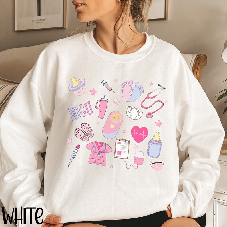May include: White sweatshirt with a pink and blue design featuring a baby in a crib, a stethoscope, a pacifier, a syringe, a heart rate monitor, a baby bottle, a thermometer, a pair of shoes, a diaper, a nurse's uniform, a clipboard, a blood bag, and a face mask. The text "NICU" is printed on the sweatshirt.