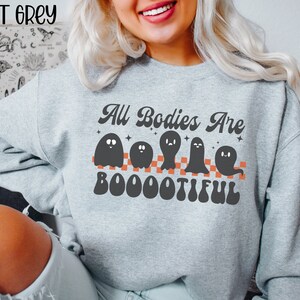May include: A grey sweatshirt with the text "All Bodies Are Boootiful" in a retro font. The text is surrounded by five black ghosts with orange checkerboard stripes.