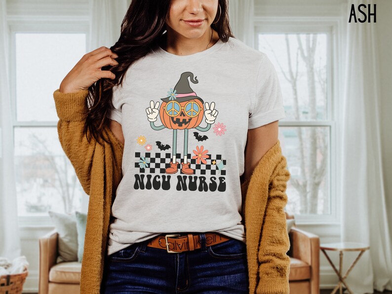 Puede incluir: Camiseta blanca con un gr&aacute;fico de una calabaza con un sombrero de bruja y gafas de sol. La calabaza est&aacute; haciendo un signo de paz y de pie sobre un patr&oacute;n a cuadros con flores. El texto "NICU NURSE" est&aacute; debajo de la calabaza.