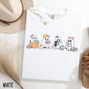 May include: White Comfort Colors t-shirt featuring a whimsical graphic of four cartoon ghosts reading books. One ghost is in a pumpkin, another has a book on its head, and the others have flowers and glasses. The word "WHITE" is at the bottom.