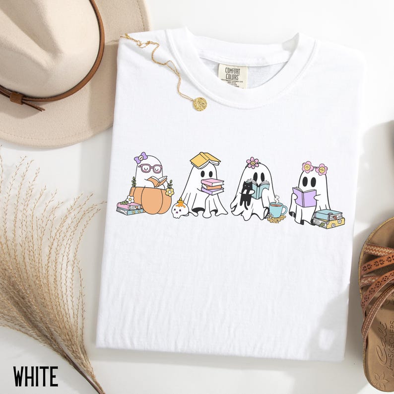 May include: White Comfort Colors t-shirt featuring a whimsical graphic of four cartoon ghosts reading books. One ghost is in a pumpkin, another has a book on its head, and the others have flowers and glasses. The word "WHITE" is at the bottom.
