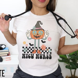 Puede incluir: Camiseta blanca con un gr&aacute;fico de una calabaza sonriente que lleva un sombrero de bruja y gafas de sol con un signo de paz. La calabaza est&aacute; levantando dos signos de paz y est&aacute; de pie sobre un patr&oacute;n a cuadros. El texto "NICH NURSE" est&aacute; impreso debajo de la calabaza.