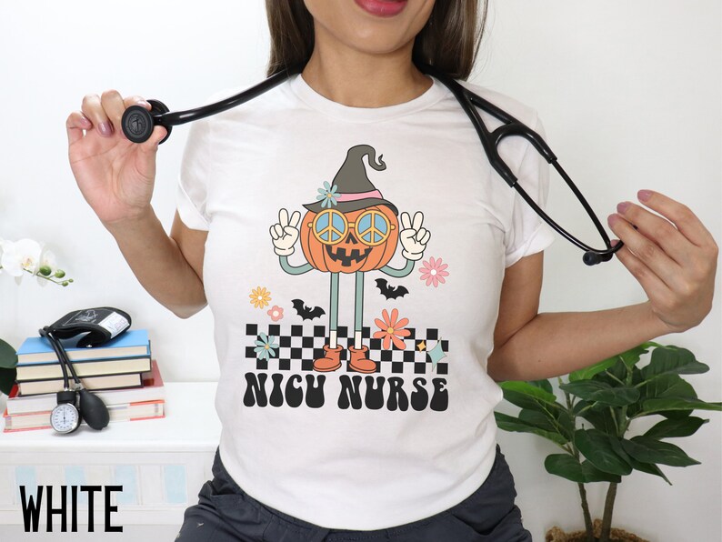 Puede incluir: Camiseta blanca con un gr&aacute;fico de una calabaza sonriente que lleva un sombrero de bruja y gafas de sol con un signo de paz. La calabaza est&aacute; levantando dos signos de paz y est&aacute; de pie sobre un patr&oacute;n a cuadros. El texto "NICH NURSE" est&aacute; impreso debajo de la calabaza.