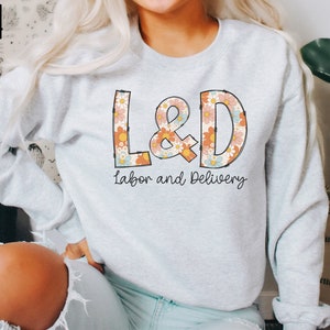 May include: Light gray sweatshirt with the text "L&D Labor and Delivery" in a retro floral design. The letters L&D are filled with a pattern of orange, pink, and blue flowers. The sweatshirt is a crew neck style.