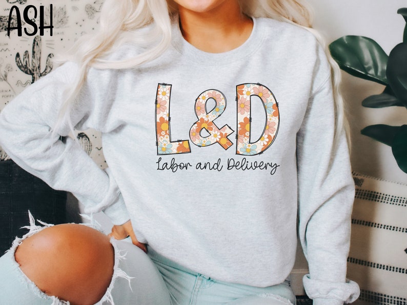 May include: Light gray sweatshirt with the text "L&D Labor and Delivery" in a retro floral design. The letters L&D are filled with a pattern of orange, pink, and blue flowers. The sweatshirt is a crew neck style.