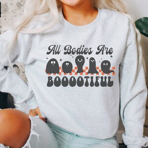 May include: A grey sweatshirt with the text "All Bodies Are Boo-o-o-tiful" in a retro font. The text is surrounded by four black ghosts with white eyes and a red and white chequered pattern.
