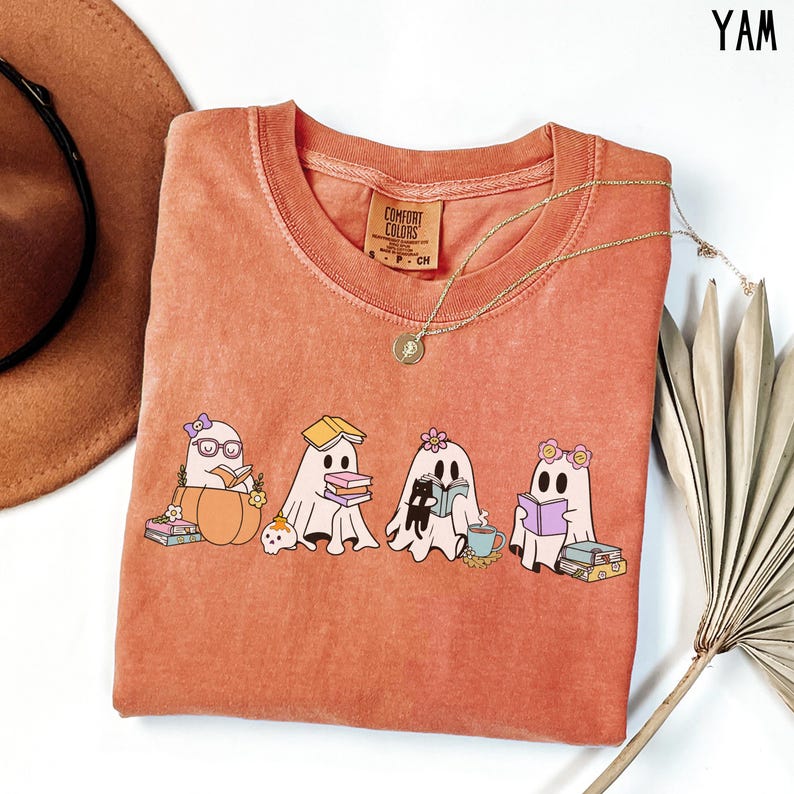 May include: A coral Comfort Colors sweatshirt featuring a graphic of four cartoon ghosts reading books. The ghosts are adorned with accessories like glasses and flowers. A gold necklace and a brown hat are also visible.