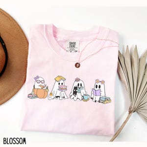 May include: A light pink Comfort Colors t-shirt featuring a graphic of four cartoon ghosts reading books. The ghosts are adorned with accessories like glasses, flowers, and a bow. The word "BLOSSOM" is printed at the bottom.