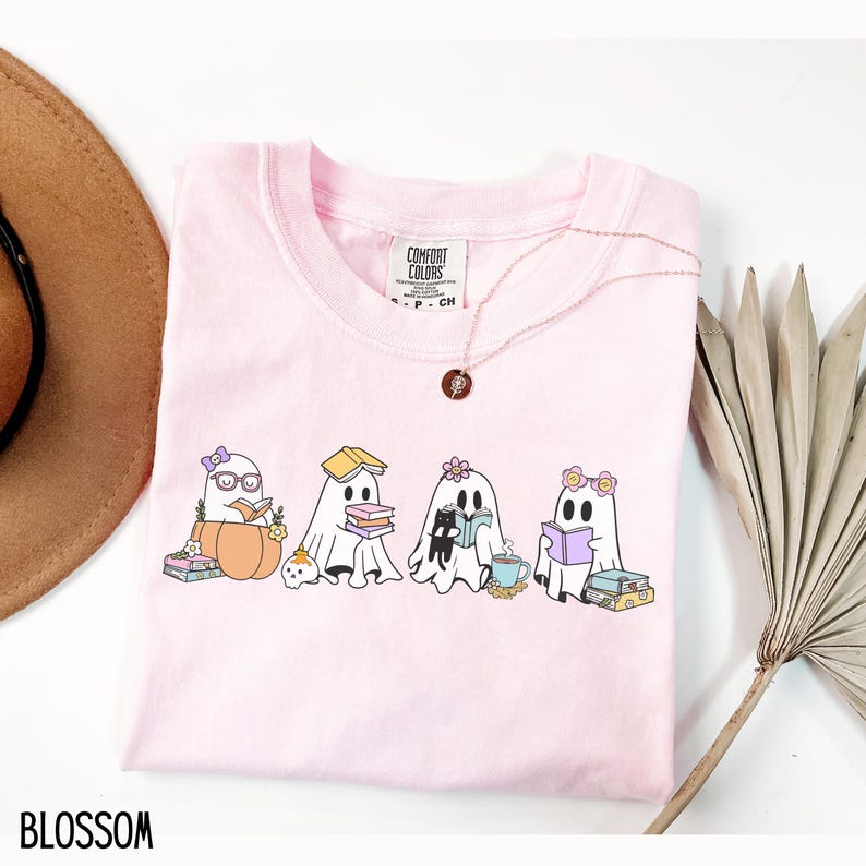 May include: A light pink Comfort Colors t-shirt featuring a graphic of four cartoon ghosts reading books. The ghosts are adorned with accessories like glasses, flowers, and a bow. The word "BLOSSOM" is printed at the bottom.